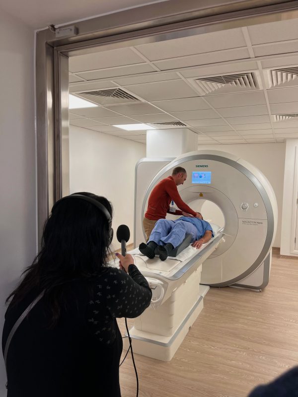 BBC producer lying down on MRI machine while healthcare professional prepares him for scan, and BBC writer and presenter records with a microphone.