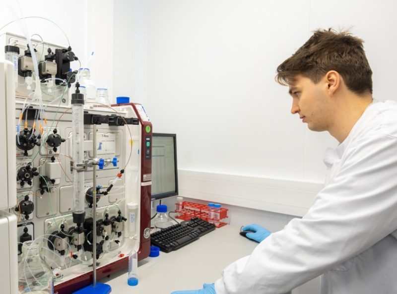 A person in a lab coat and blue gloves look at a piece of scientific equipment and computer.